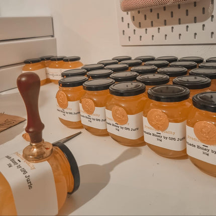 Jars of honey with black lids on a white surface, with a person wearing pink shoes in the background.