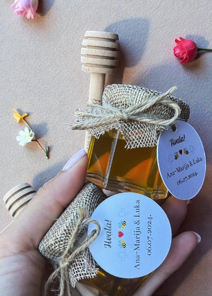 Small bottle of honey with a wooden dipper, tied with burlap ribbon, held by a hand on a light background with flowers.